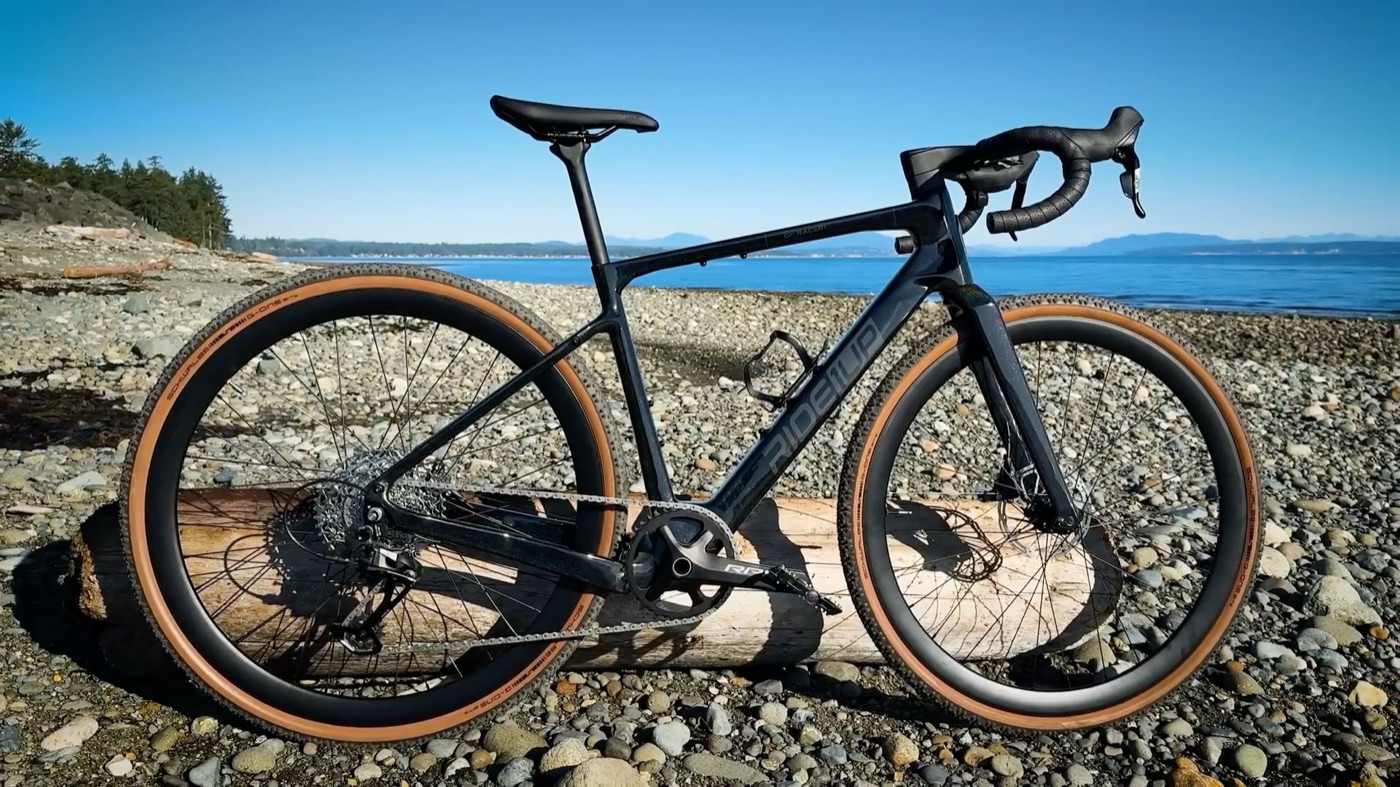 Ride1Up CF Racer1 parked on a pebble beach with the ocean behind it